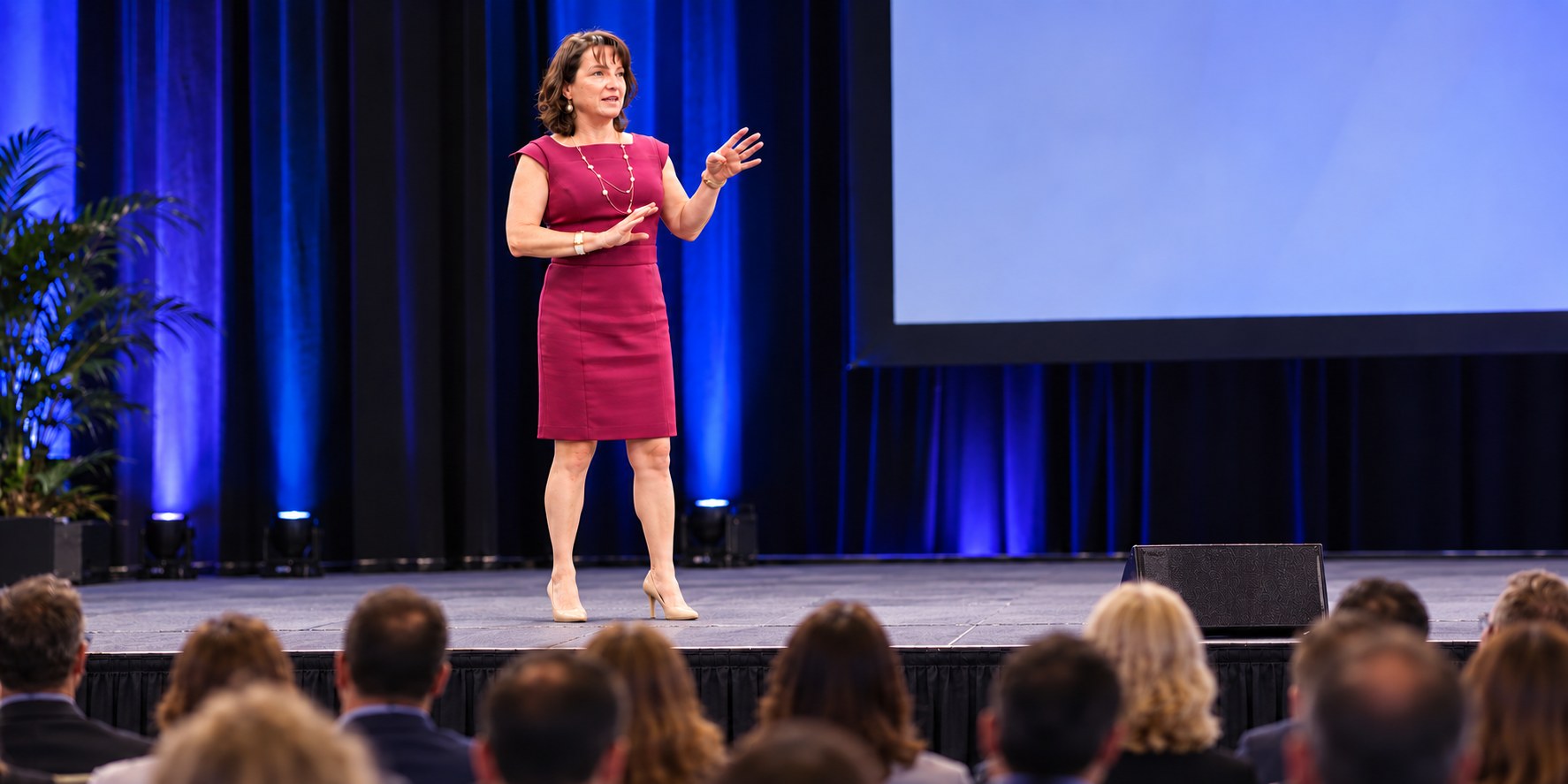 Anna Conrad presenting on a blue-lit stage, gesturing toward a seated conference audience
