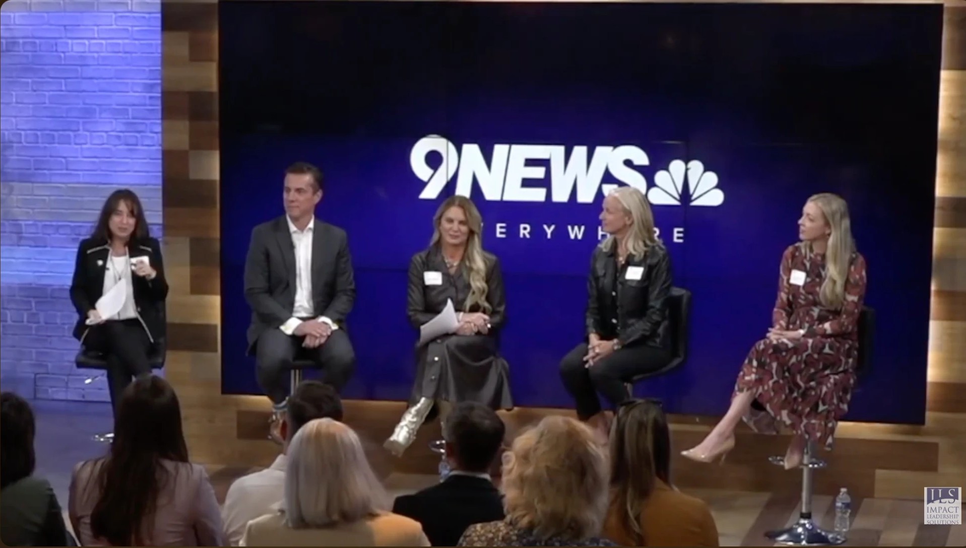 Five Executive Voices panelists seated on a stage before an audience, with a 9NEWS screen behind them